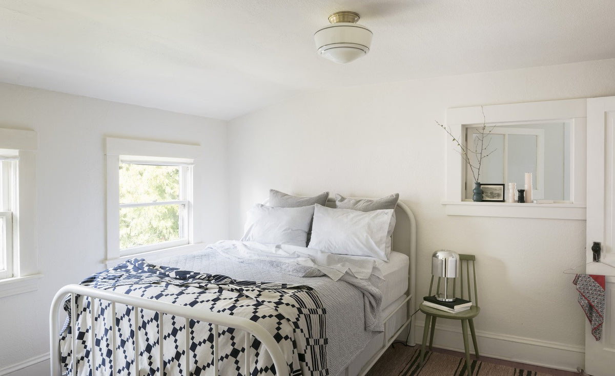 Sunny bedroom with white metal bed and patterned quilt, flush-mount ceiling light and green chair holding a bedside lamp