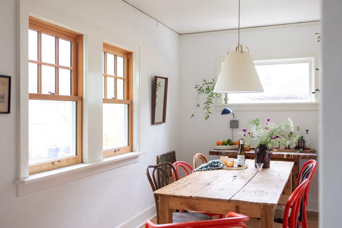 Dining room with pleated white pendant light above a rustic wooden table, red bentwood chairs, and a vase of wildflowers