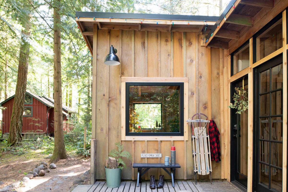 Sunlit wooden cabin porch with bench, boots, sled and red plaid jacket by glass doors in a forest; sign reads Please remove shoes