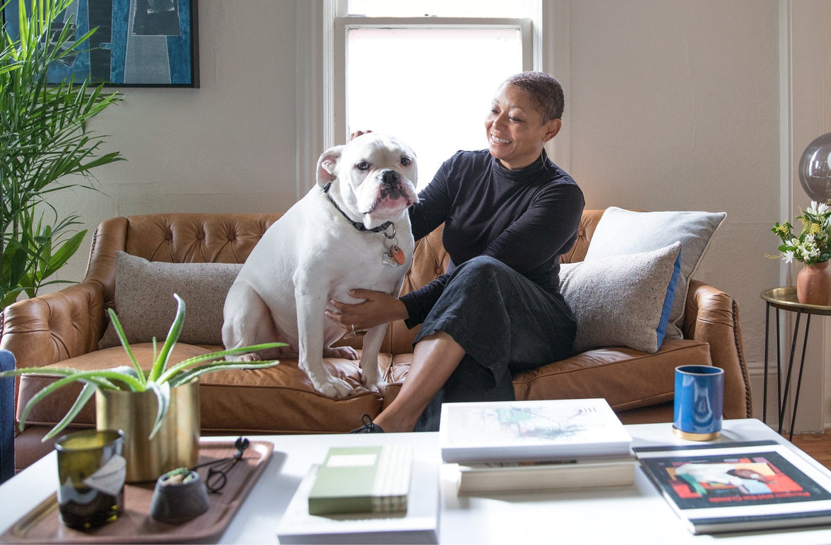 Woman smiling and petting a white bulldog on a tan leather sofa in a sunlit living room with coffee table and plants