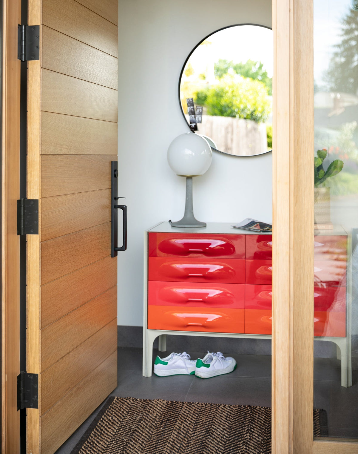 Entryway with an open wooden door, red ombré chest of drawers, globe table lamp and round wall mirror reflecting greenery