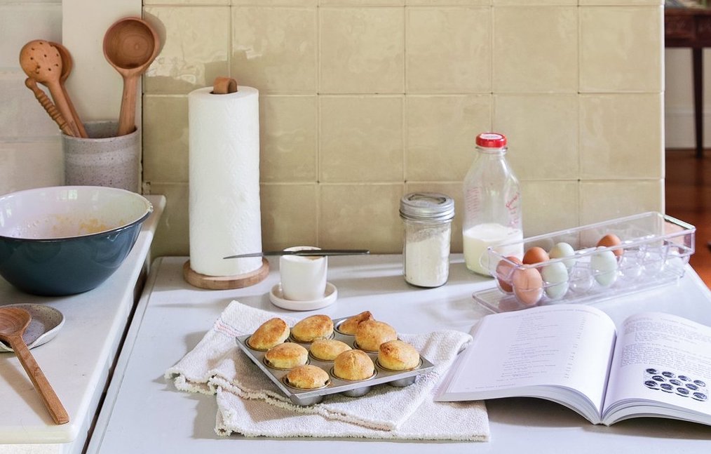 Custardy popovers cooling in a tin on a kitchen counter with mixing bowl, milk bottle, eggs, and open cookbook