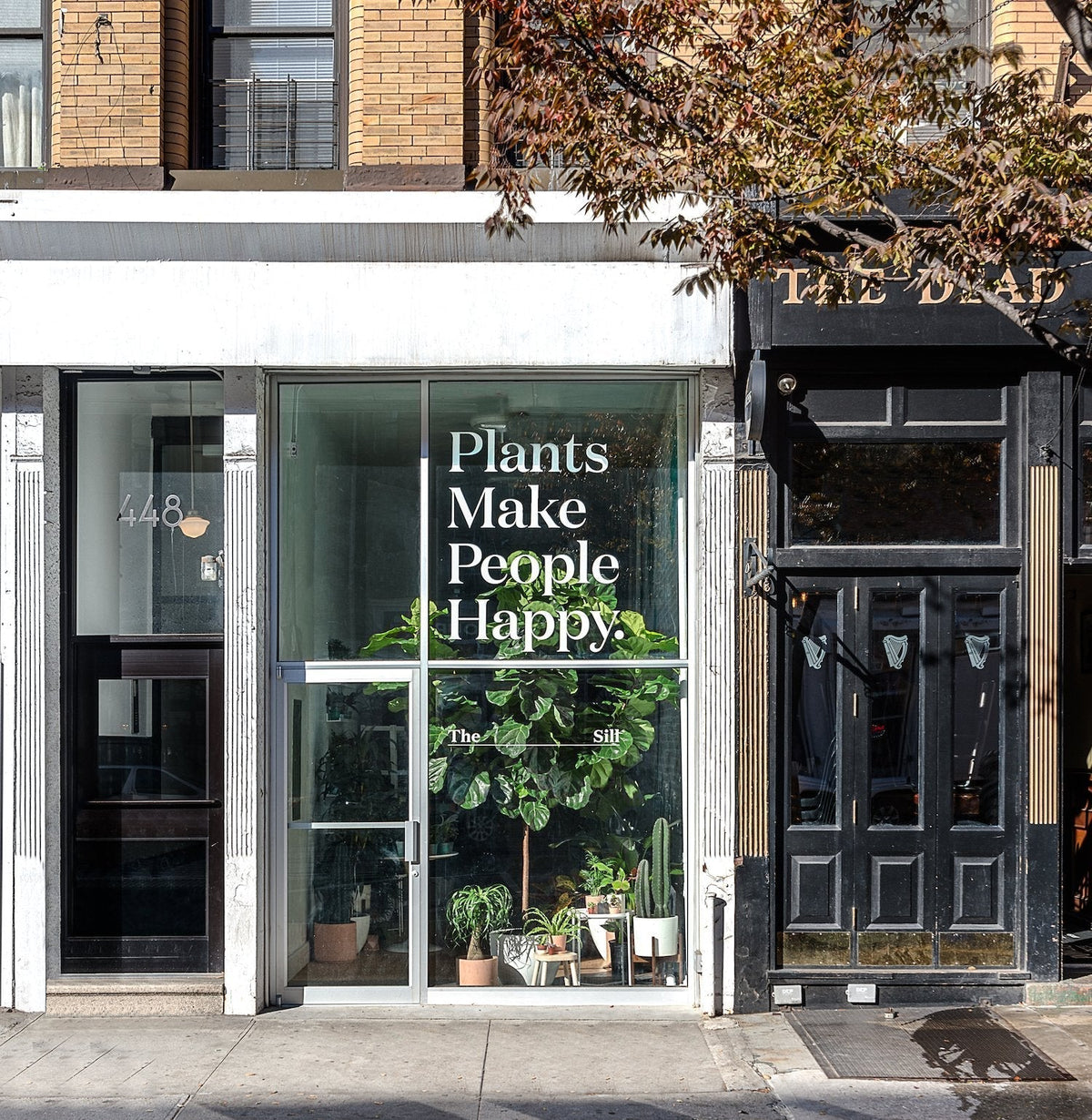 Storefront window of a plant shop with potted houseplants and a large sign reading ‘Plants Make People Happy’ on a city sidewalk
