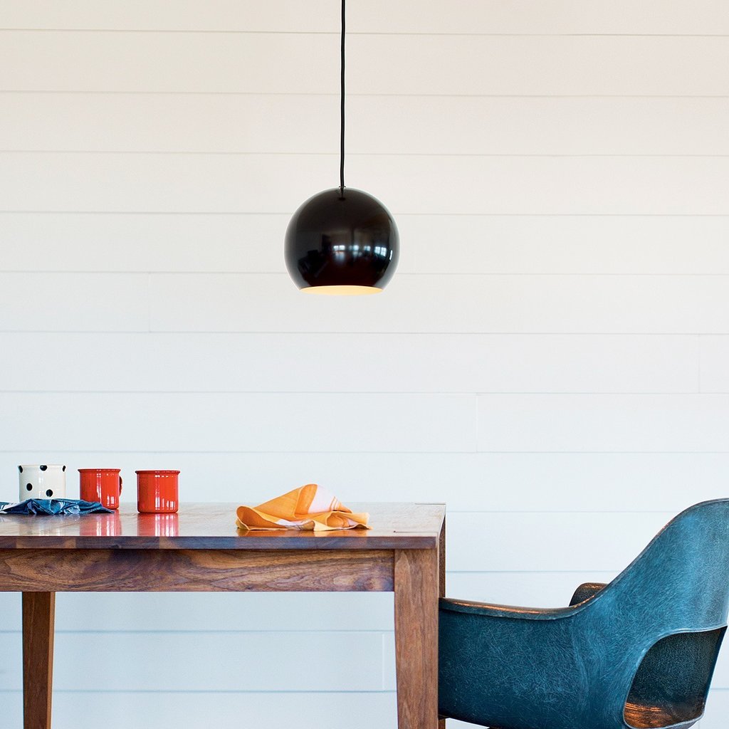 Black globe pendant light over dining table with teal chair, red mugs, and orange napkin against white shiplap wall