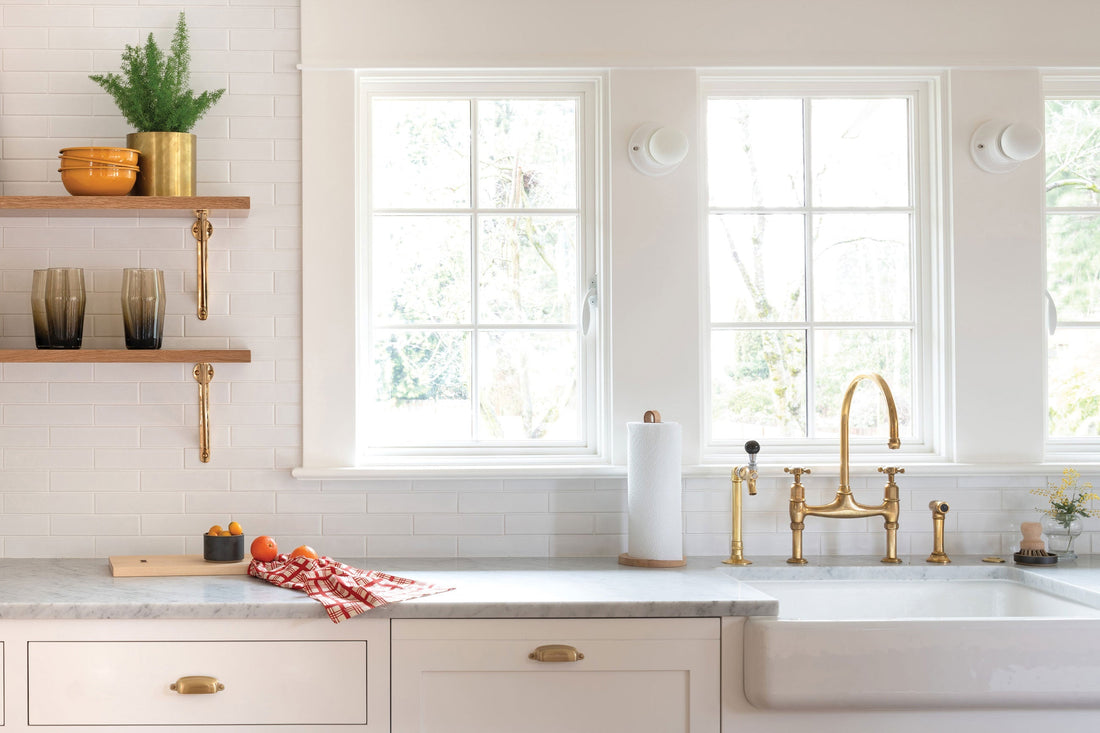 Brass faucet and cabinet handles in a bright white kitchen with marble countertop, farmhouse sink, and open wood shelves