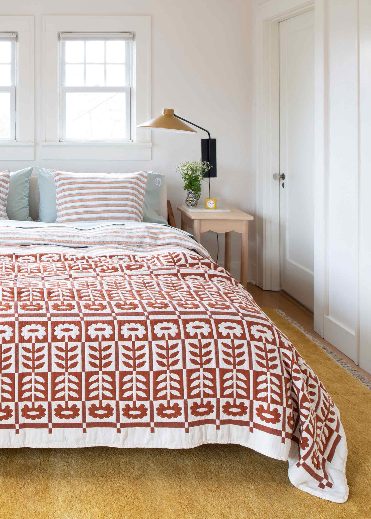 Bed with rust-and-white block-print quilt and striped pillows in a bright white bedroom with wall-mounted lamp and nightstand