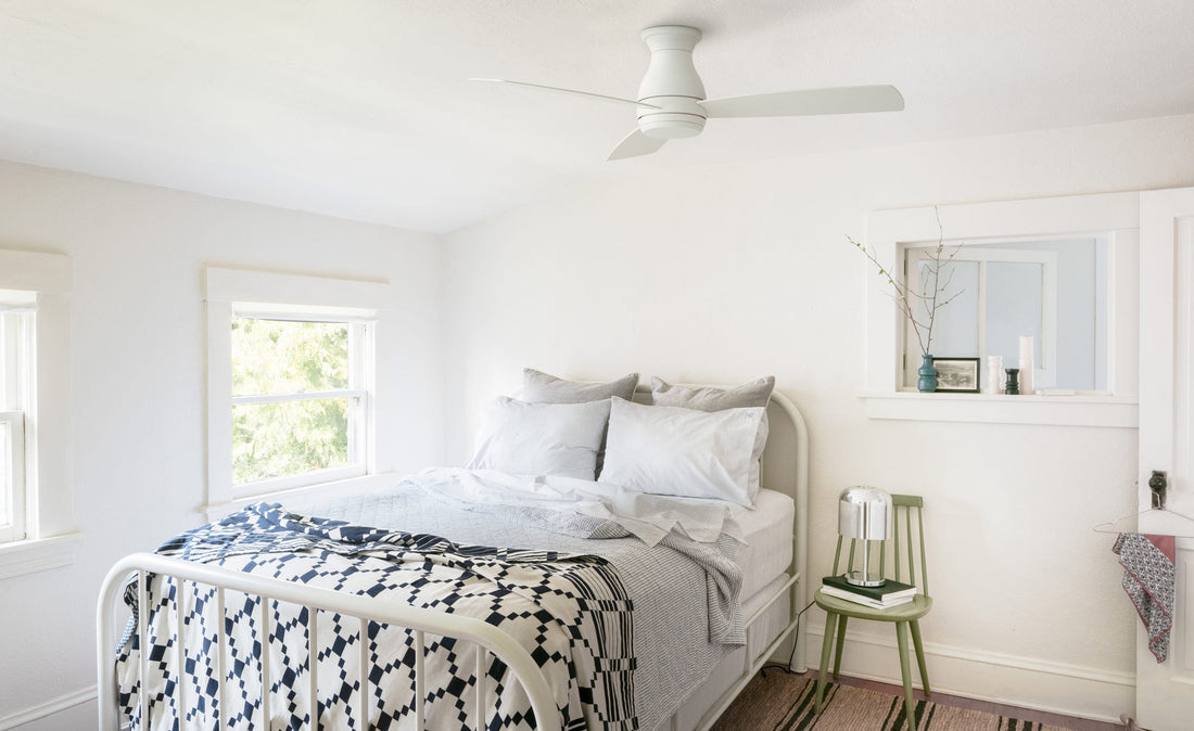 White ceiling fan above a sunlit small bedroom with a white metal bed, blue patterned quilt, and green side chair