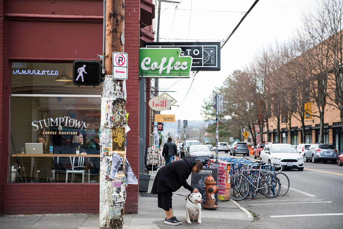Woman petting a large white dog outside Stumptown Coffee Roasters beneath a green ‘Coffee’ sign on a Portland street