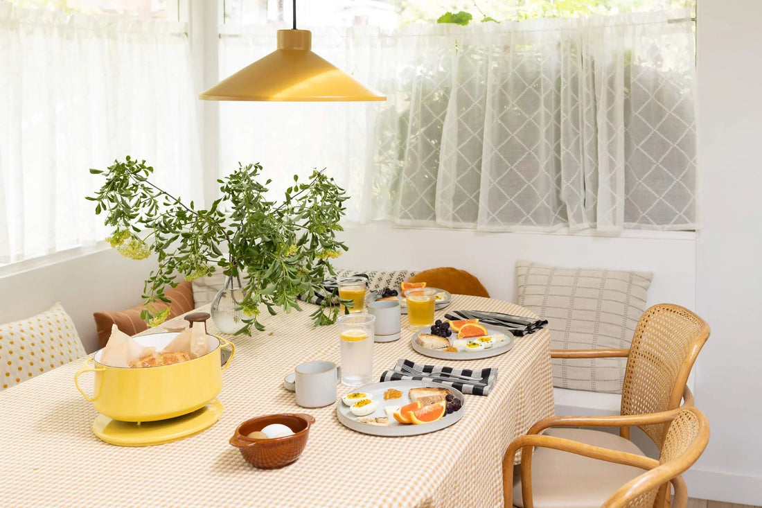 Spring brunch table set with gingham tablecloth, yellow pendant light, plates of citrus and eggs, glasses, wicker chairs, and a vase of greenery