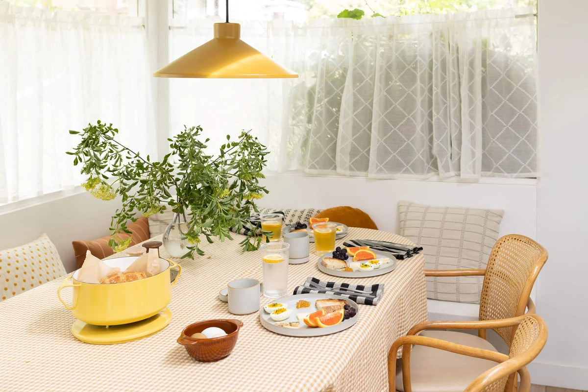 Spring brunch table set with gingham tablecloth, yellow pendant light, plates of citrus and eggs, glasses, wicker chairs, and a vase of greenery