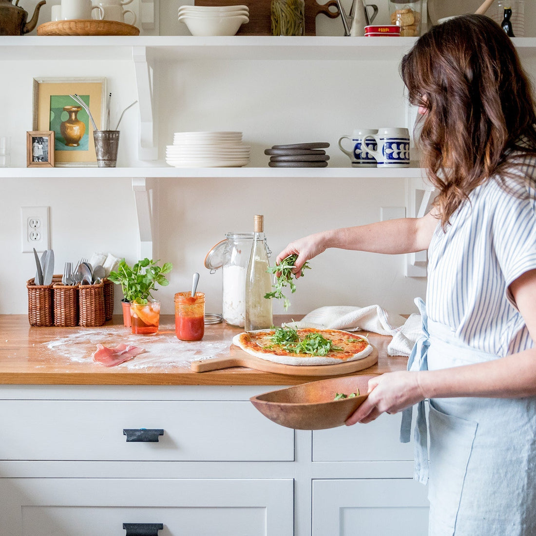 Woman sprinkling arugula over a homemade pizza on a wooden countertop in a bright kitchen with open shelves