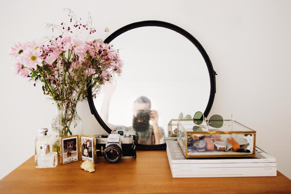 Round mirror on wooden dresser with pink wildflowers in a glass vase, vintage Canon camera, framed photos, glass jewelry box with watches and sunglasses, photographer reflected in the mirror