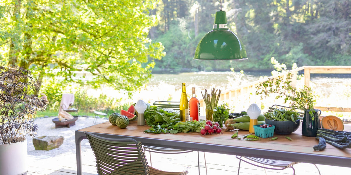 Outdoor dining table arranged with fresh summer vegetables, fruit, and drinks under a green pendant lamp on a lakeside deck