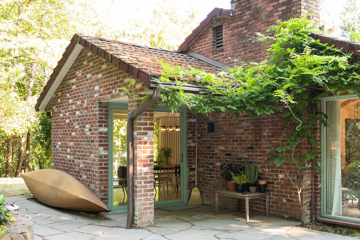 Brick cottage exterior with green-framed glass doors, climbing vines over the roof, a canoe on the stone patio and potted plants on a bench