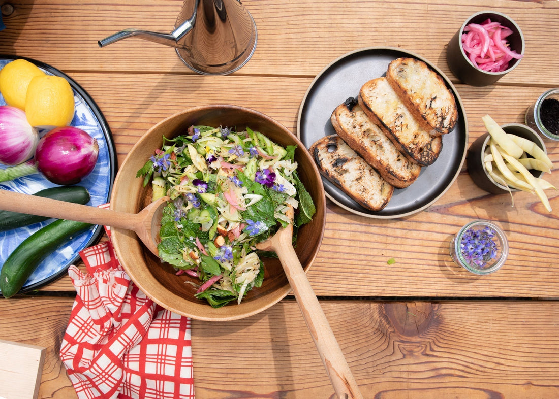 Overhead view of a wax bean and herb salad in a wooden bowl with wooden serving spoons, grilled sourdough slices, pickled onions and lemons on a wooden table