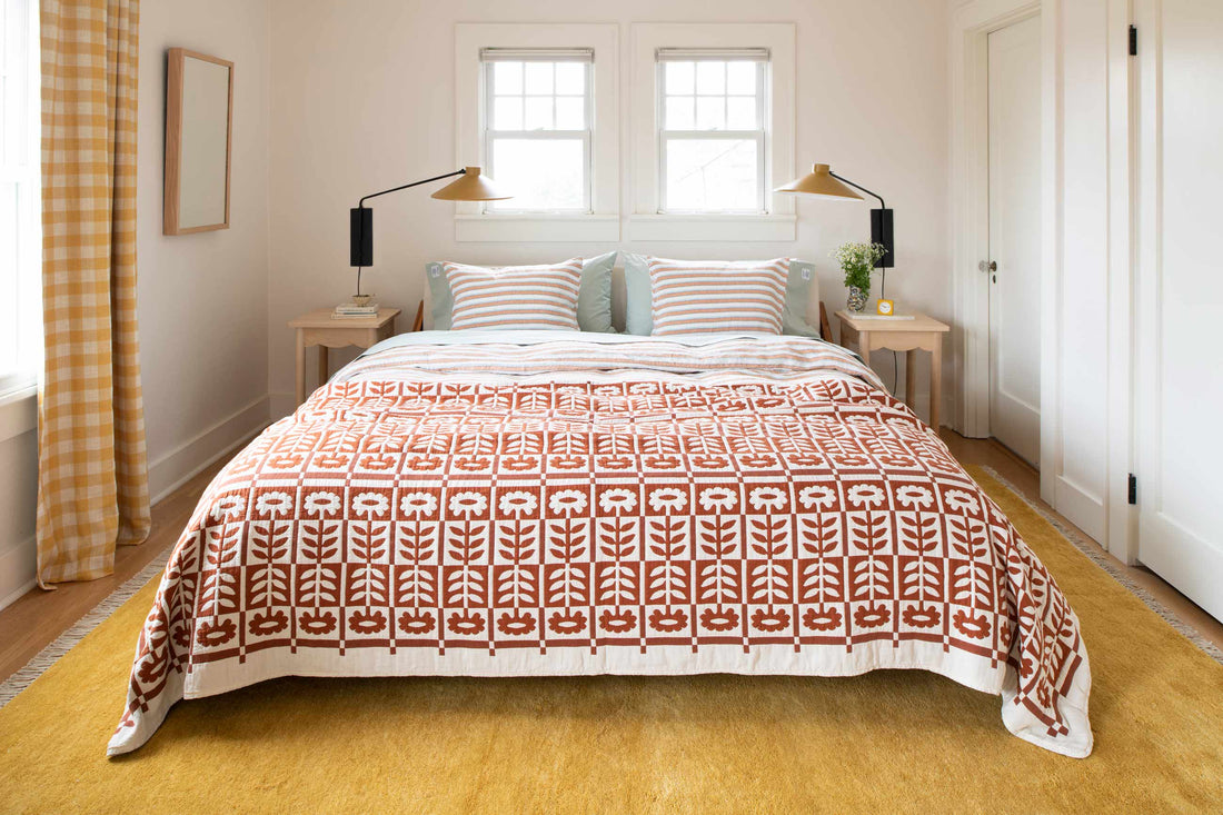 Bed with rust-and-white floral quilt and striped pillows in a bright bedroom with yellow rug and gingham curtains