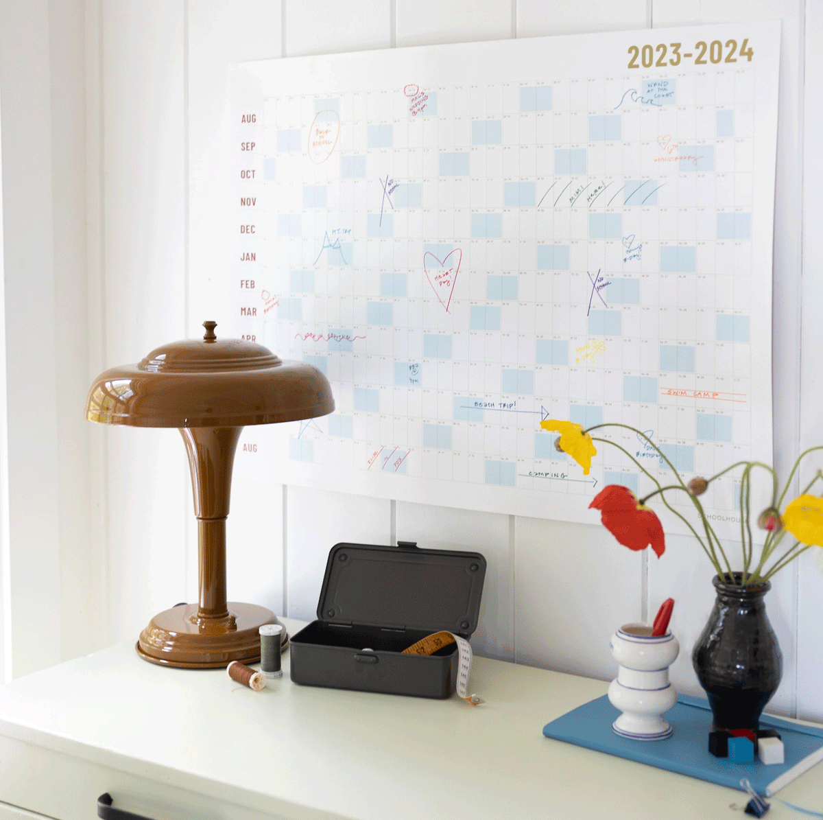 2023-2024 academic wall calendar hung above a desk, marked with colorful handwritten notes, with a brown table lamp and a vase of flowers