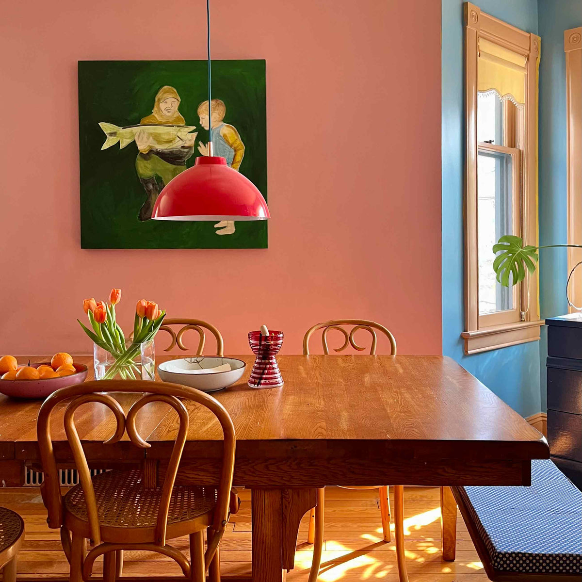 Dining table arranged with tulips, a bowl of oranges, and a red pendant light in a colorful dining room with pink walls and blue window trim