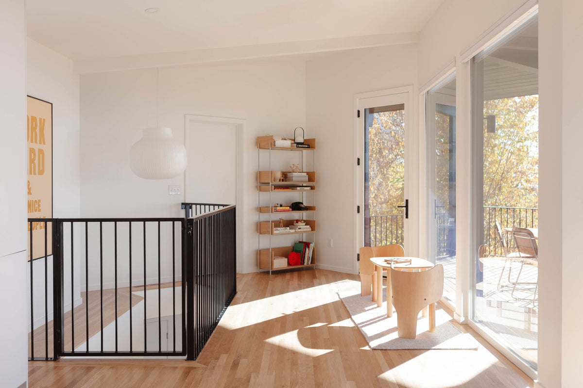 Sunlit upstairs landing with black metal stair railing, a wooden children’s table and chairs on a small rug beside sliding glass doors to a balcony