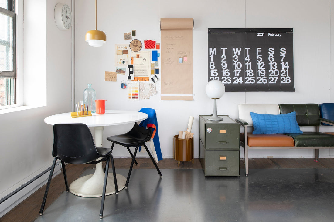 Sunlit studio seating area with round white table and black chairs, bulletin board, February 2021 wall calendar, metal filing cabinet and bench