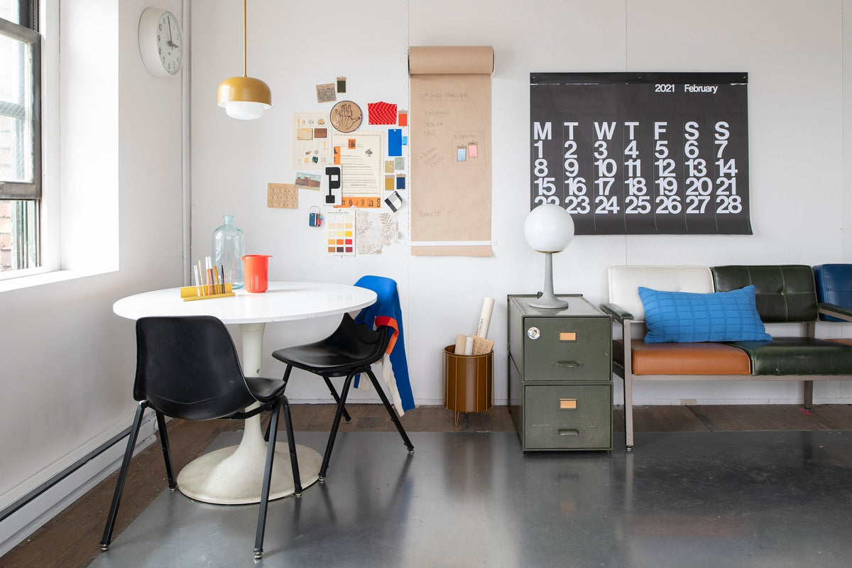 Sunlit studio seating area with round white table and black chairs, bulletin board, February 2021 wall calendar, metal filing cabinet and bench