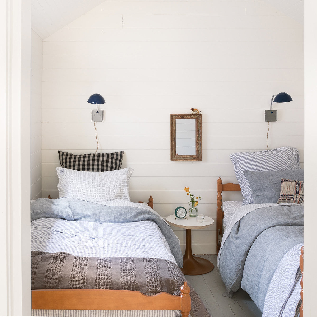 Two twin beds dressed in diamond-ticking quilts and linen shams in a bright white paneled bedroom with a small round bedside table and navy wall lamps