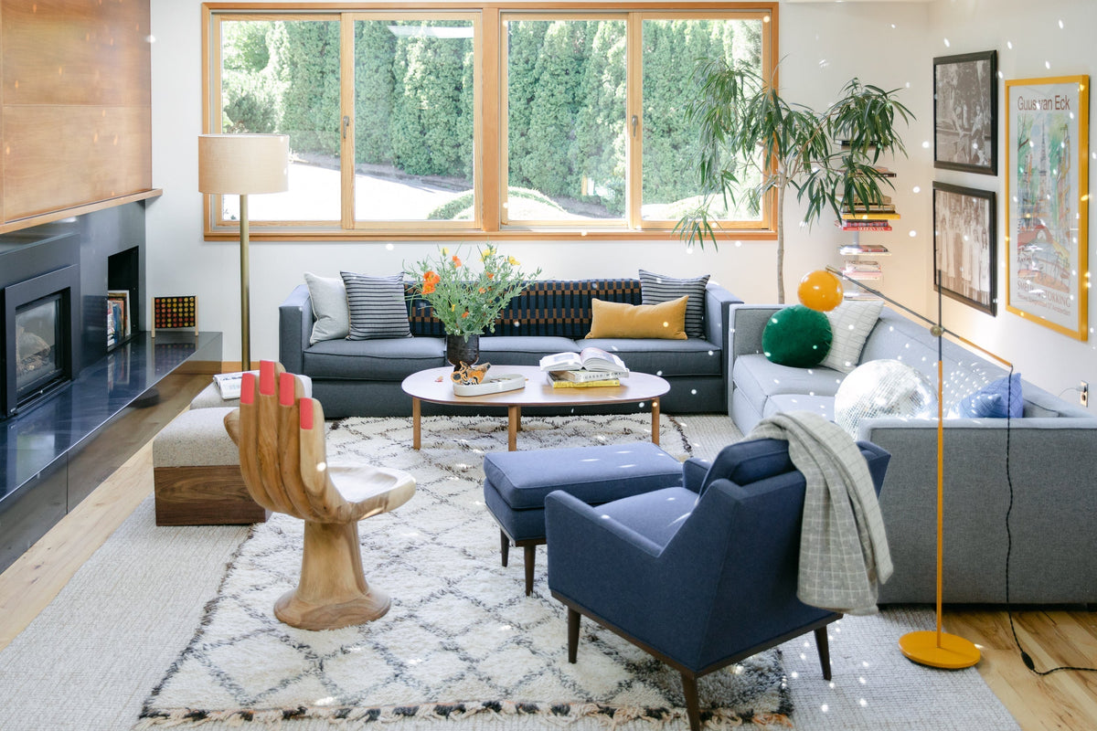 Bright living room with a gray L-shaped sofa, blue armchair and ottoman, wooden hand-shaped chair, round coffee table, patterned rug, and large window showing hedges