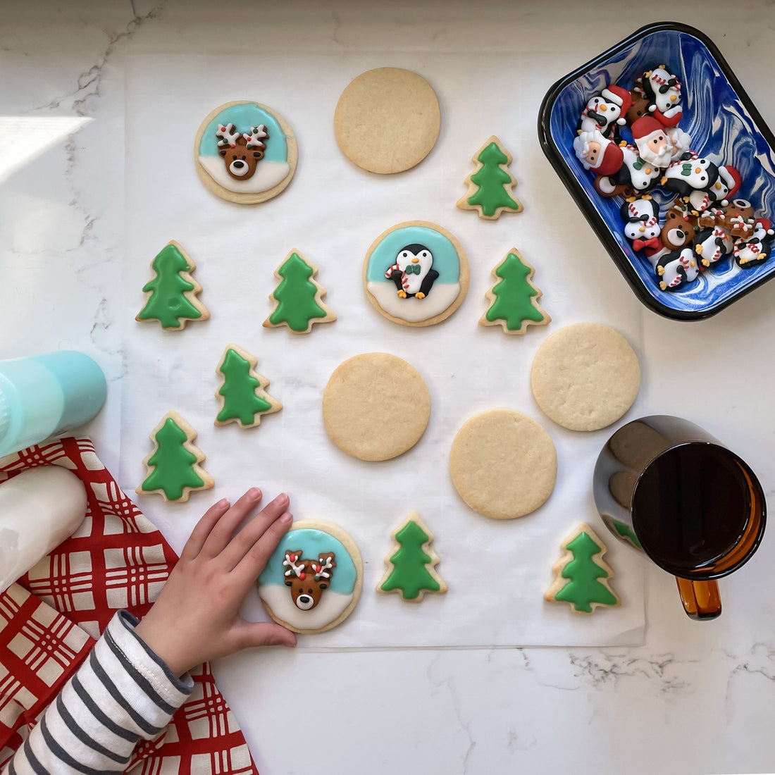 Child’s hand reaching for decorated sugar cookies on parchment with green tree cookies and round reindeer and penguin cookies