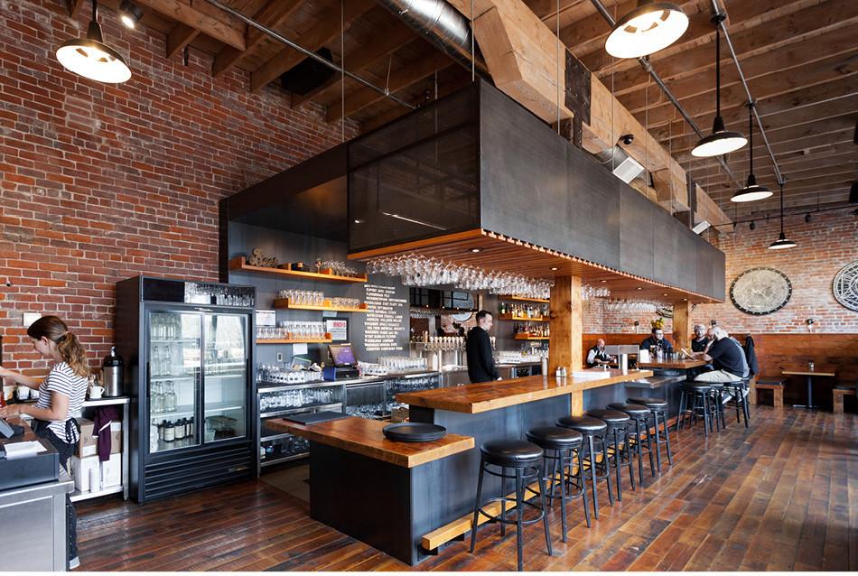 Taproom interior with long wooden bar, black stools, hanging glassware, exposed brick walls, and patrons seated at the bar