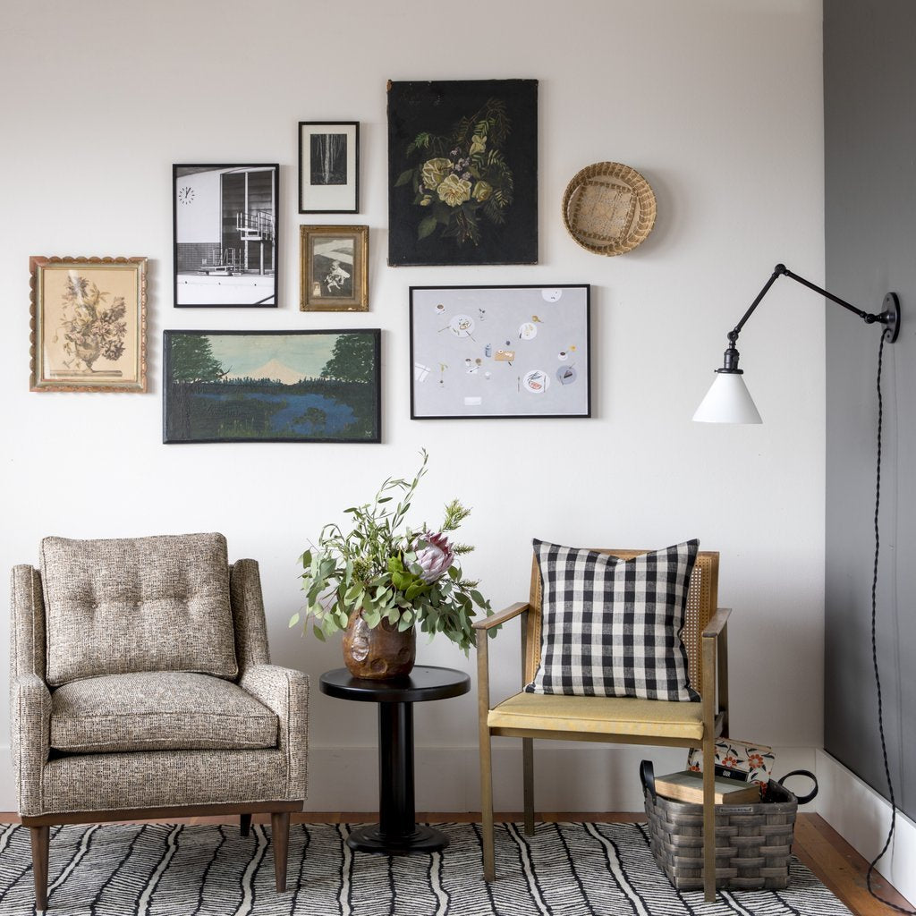 Two chairs flanking a round black side table with a vase of greenery under a gallery wall and a white swing-arm lamp