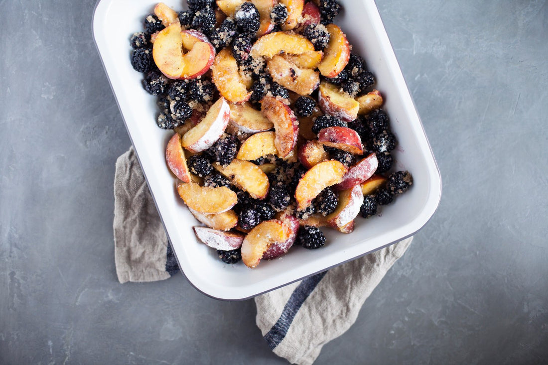 Overhead view of a white baking dish filled with sliced peaches and blackberries tossed with sugar on a gray countertop with a linen towel