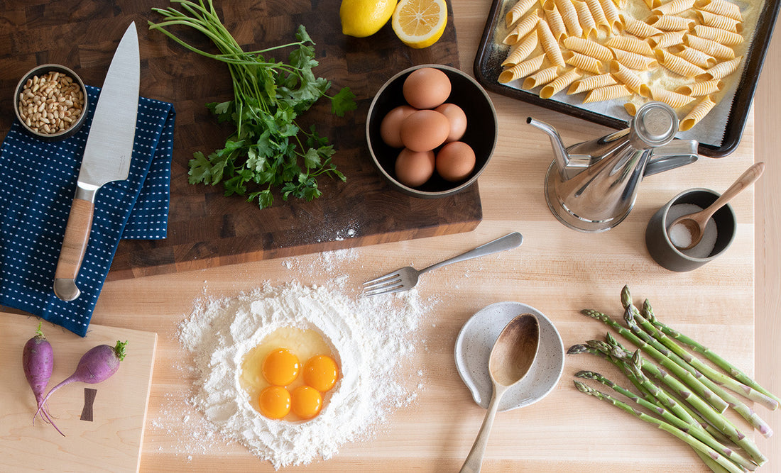 Overhead view of kitchen prep for spring pasta with eggs in a flour well, asparagus, parsley, pasta sheets, knife, and oil
