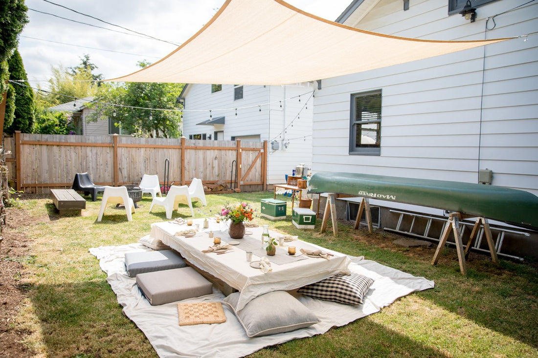 Low picnic table on blankets with cushions and place settings under a triangular shade sail in a sunny backyard
