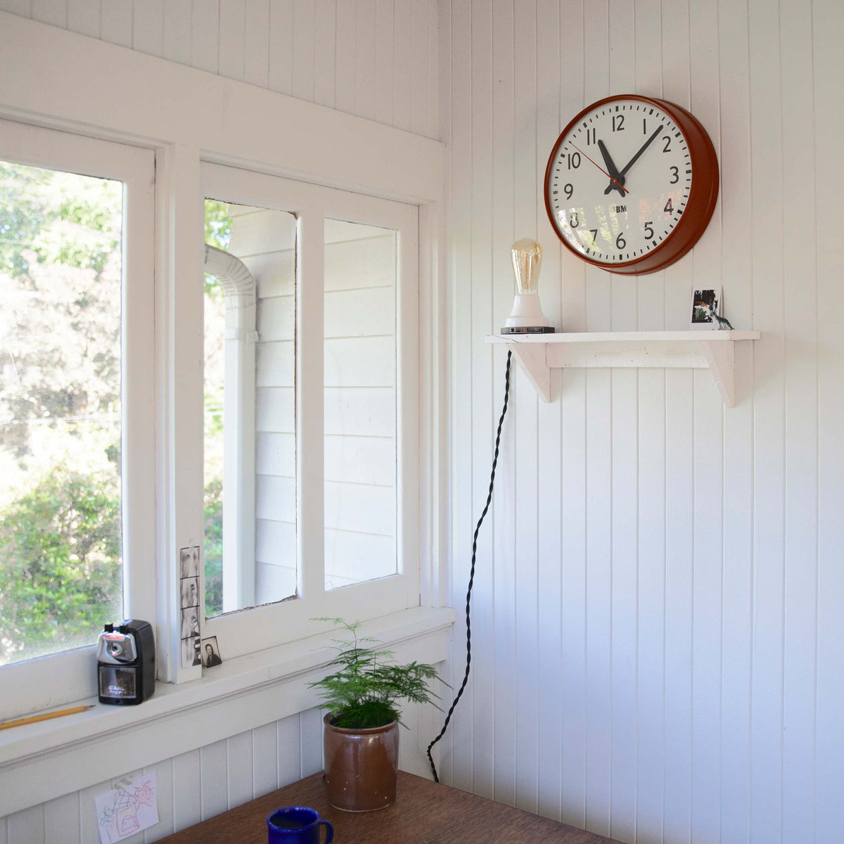 IBM wall clock with red metal frame hanging above a small white shelf on a white beadboard wall beside a sunlit window