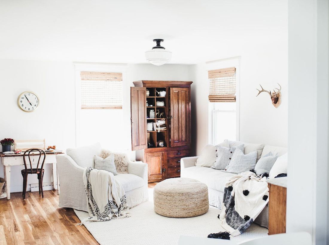 Bright living room with two white slipcovered sofas, round woven ottoman, open wooden cabinet, sunlit windows, and neutral textiles