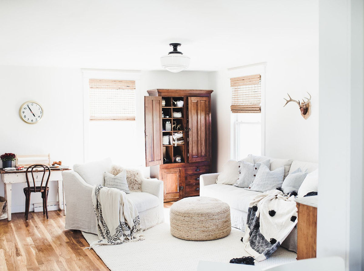 Bright living room with two white slipcovered sofas, round woven ottoman, open wooden cabinet, sunlit windows, and neutral textiles