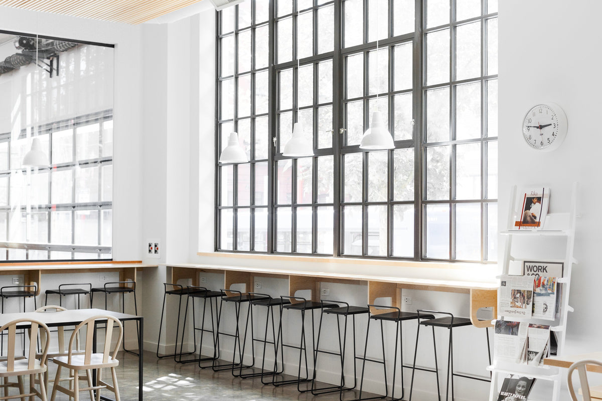 Row of stools at a sunlit counter in a minimalist coworking studio with tall black grid windows and white pendant lights