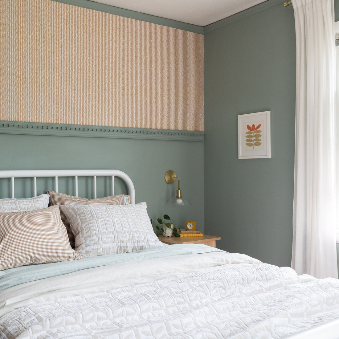 White metal bed with layered white quilt and beige gingham pillows in a sage green bedroom with brass sconce and nightstand
