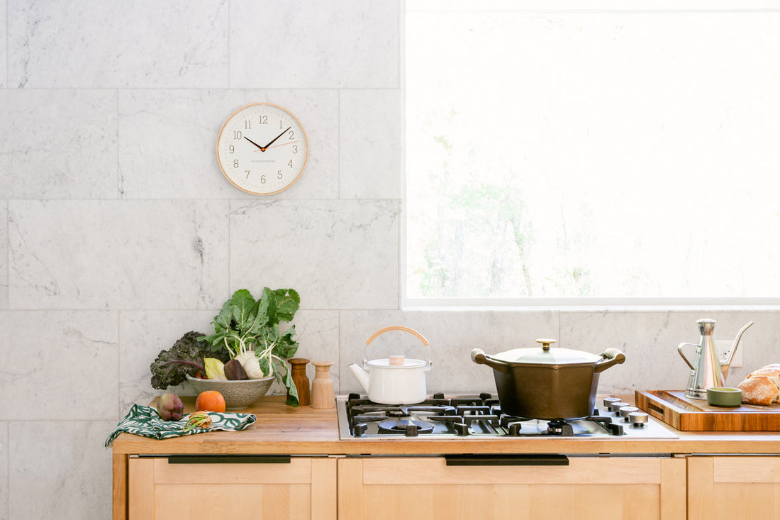 Bright kitchen showing a black pot simmering on a gas stove, a kettle, vegetables and bread on a wooden counter, wall clock
