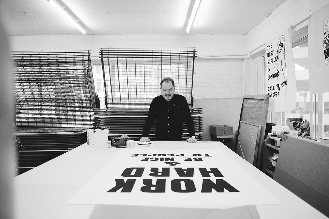 Artist Anthony Burrill leaning over a work table in his studio with a large poster reading “WORK HARD & BE NICE TO PEOPLE”