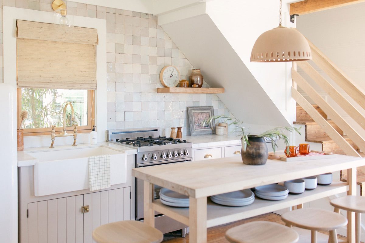 Sunlit farmhouse kitchen with a wooden island and stools, apron-front sink with brass faucet, white tiled backsplash, and pink pendant light