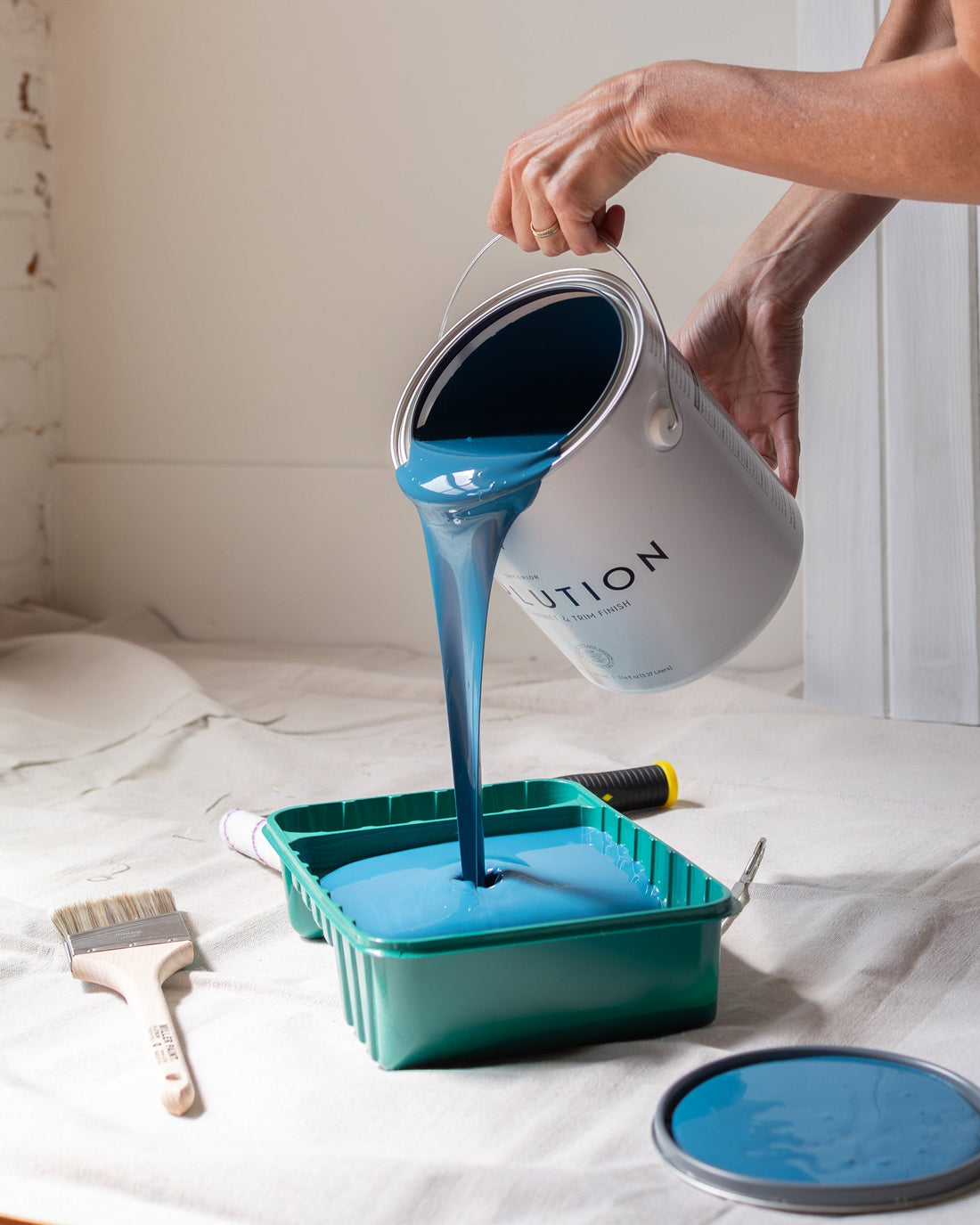 Hands pouring blue paint from a can into a roller tray on a drop cloth