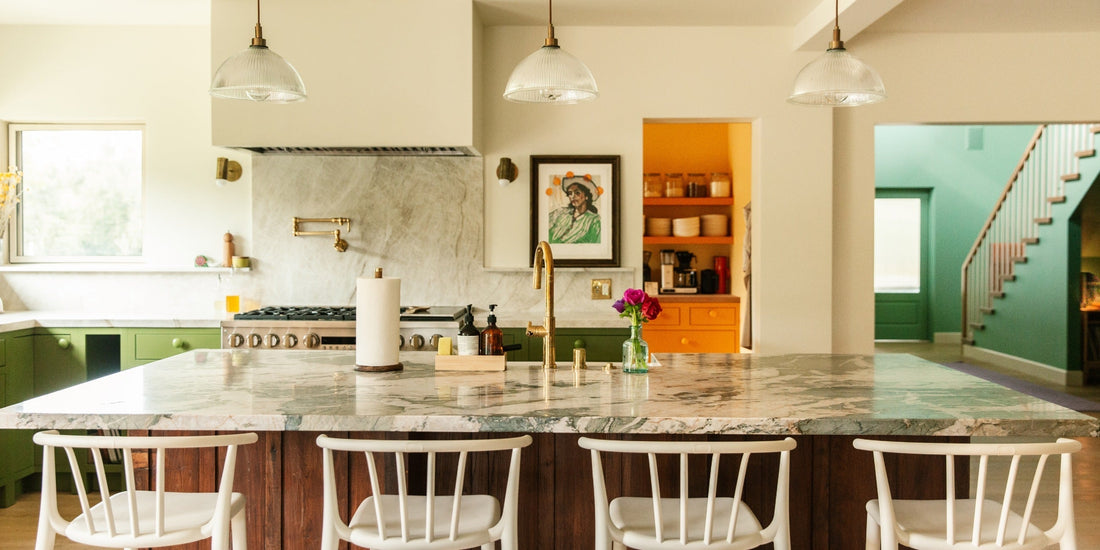 Sunlit farmhouse kitchen with marble island, brass faucet, white stools, green cabinets, and an open orange pantry