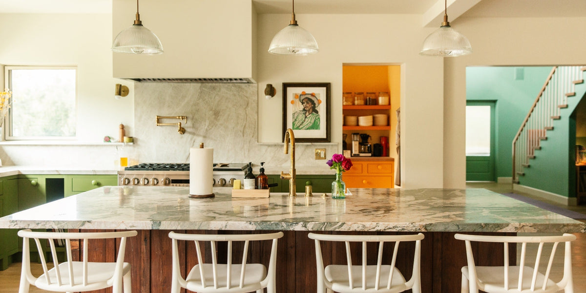 Sunlit farmhouse kitchen with marble island, brass faucet, white stools, green cabinets, and an open orange pantry