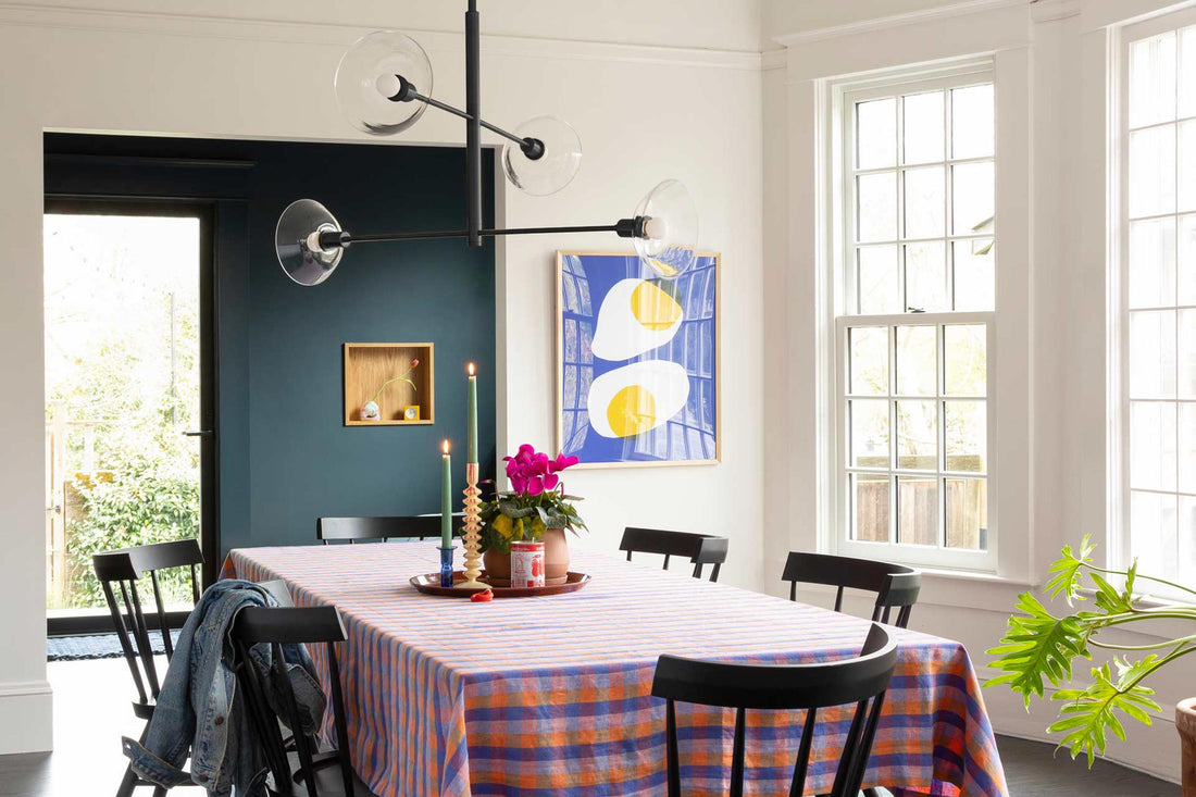 Dining room with framed sunny-side-up print, long table with blue-orange checked cloth, black chairs, glass pendant light