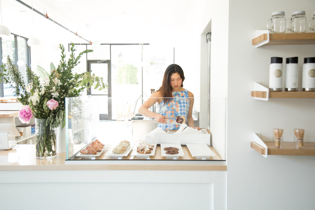 Woman packing a cookie into a takeaway box behind a glass pastry case at a bright, minimalist tea bar
