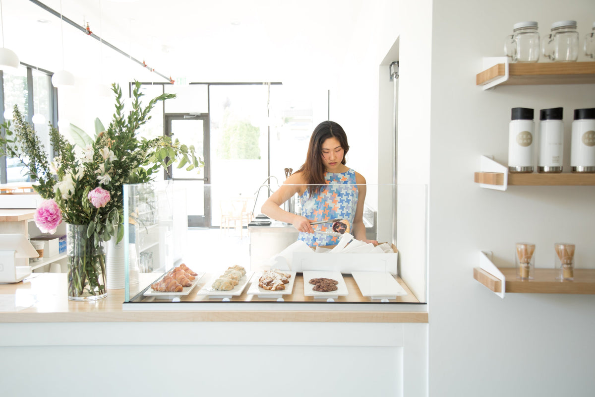 Woman packing a cookie into a takeaway box behind a glass pastry case at a bright, minimalist tea bar