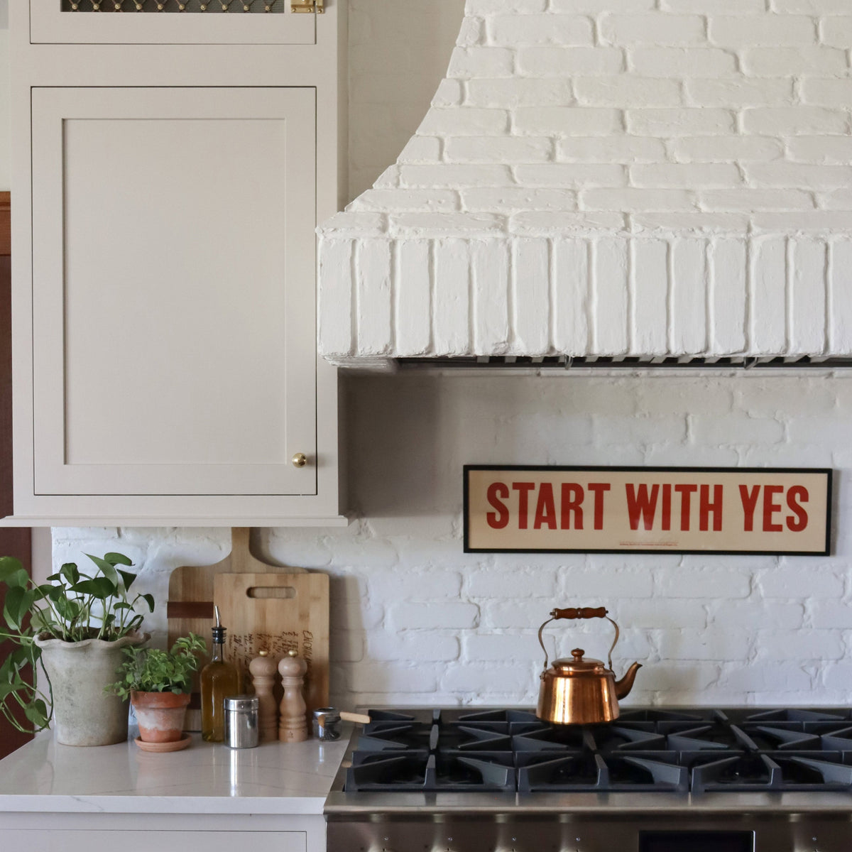 Kitchen range with painted white brick hood, copper kettle on the stove, potted herbs and cutting boards on the counter, and a framed ‘START WITH YES’ sign on the wall