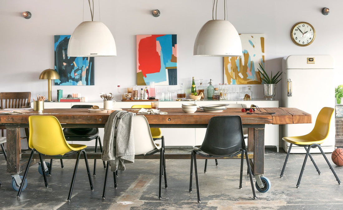 Dining room with two white pendant lights above a rustic wood table, mixed yellow and black chairs, abstract wall art, and a vintage fridge