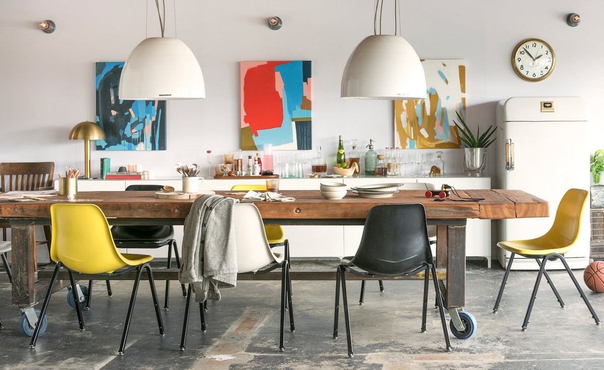 Dining room with two white pendant lights above a rustic wood table, mixed yellow and black chairs, abstract wall art, and a vintage fridge